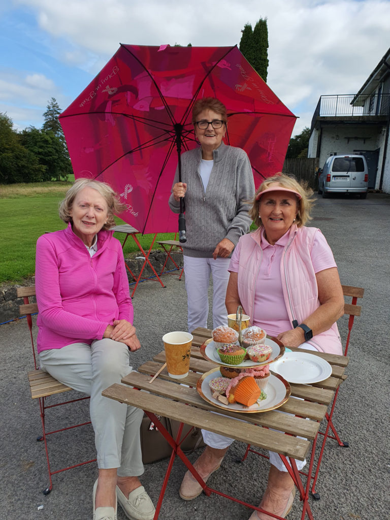 Three women enjoying tea and cupcakes outdoors, with a large pink umbrella, smiling at the camera.