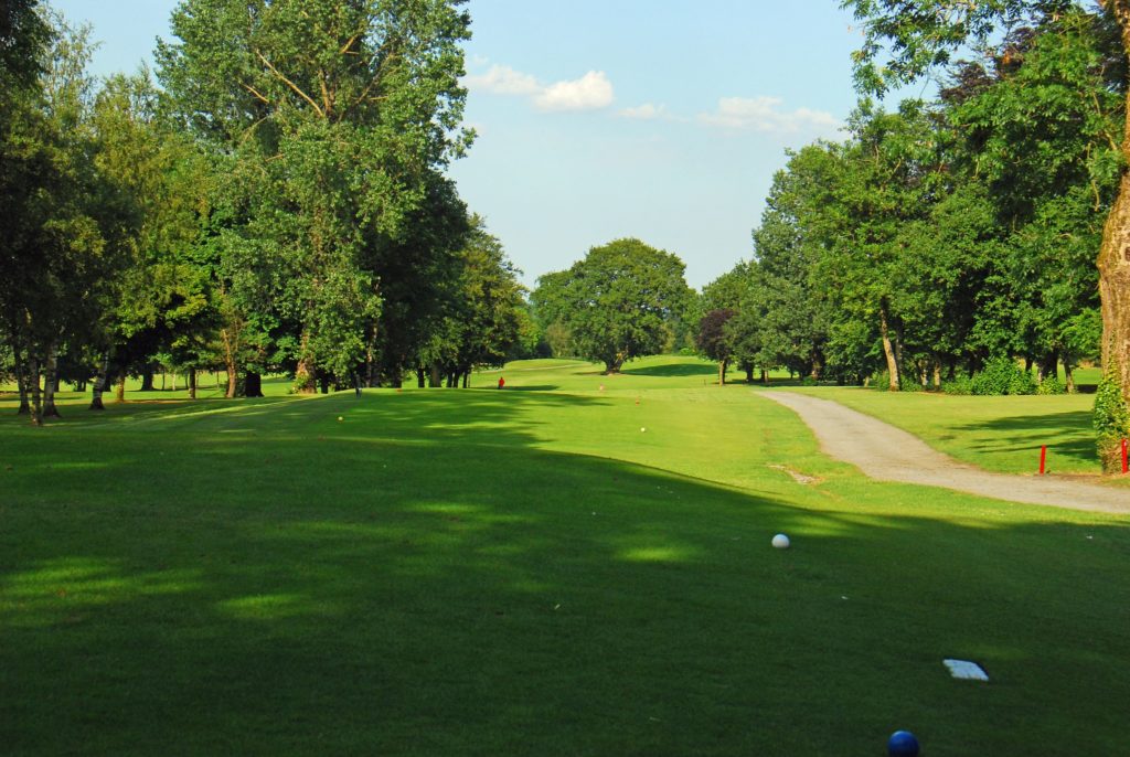 Sunny golf course view with lush green fairway, lined with trees, and a gravel path on the right.