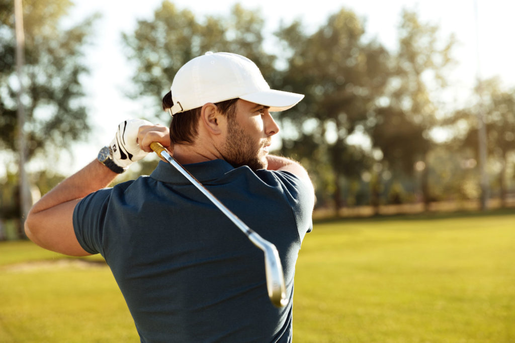 Golfer in white cap and blue shirt swinging club on sunny golf course.