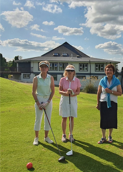 Three women standing on a golf course, sunny day, clubhouse in background.