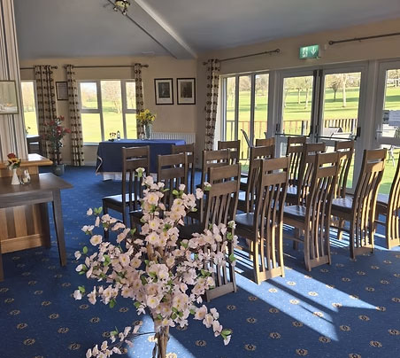 Sunlit room with wooden chairs and floral decor, overlooking a golf course through large windows. Ideal for events or meetings.