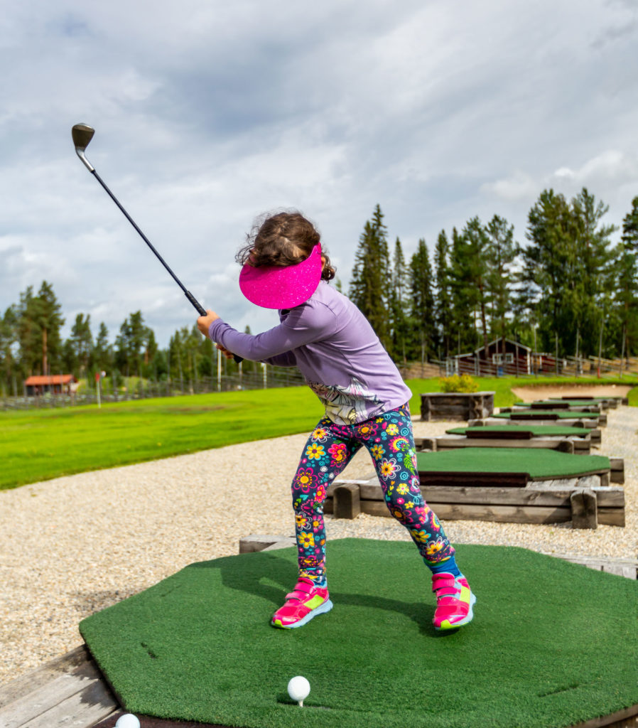 Child practicing golf swing on a driving range, wearing colorful outfit and pink visor.