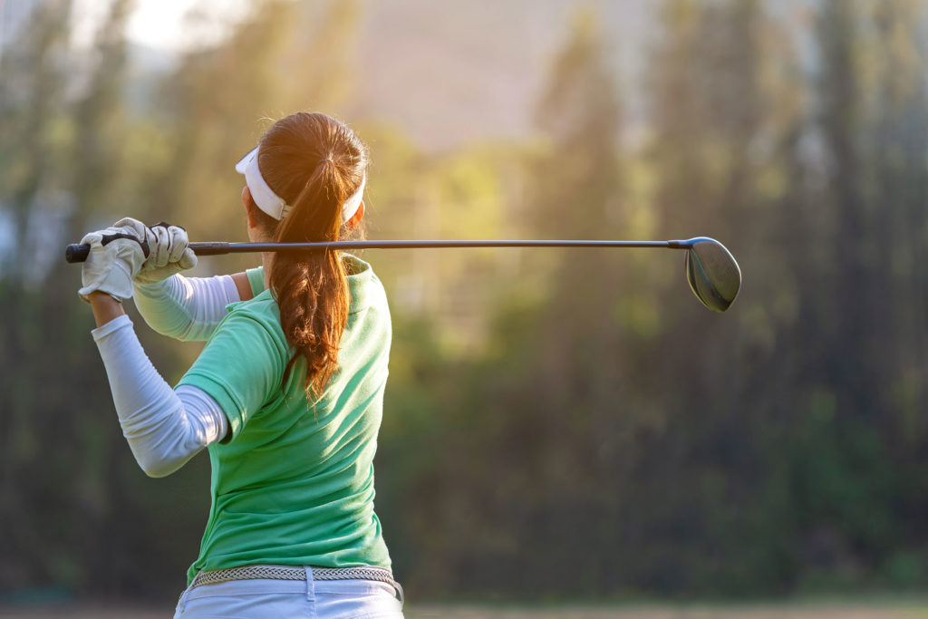 Golfer in green shirt and visor swinging club on sunny course with trees in background.