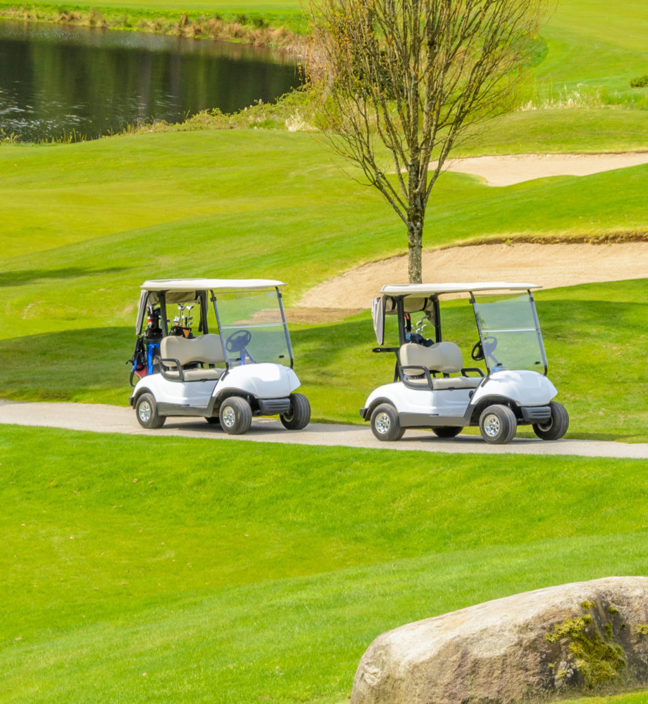 Golf carts parked on lush green course beside a pond and sand bunker under a clear sky.