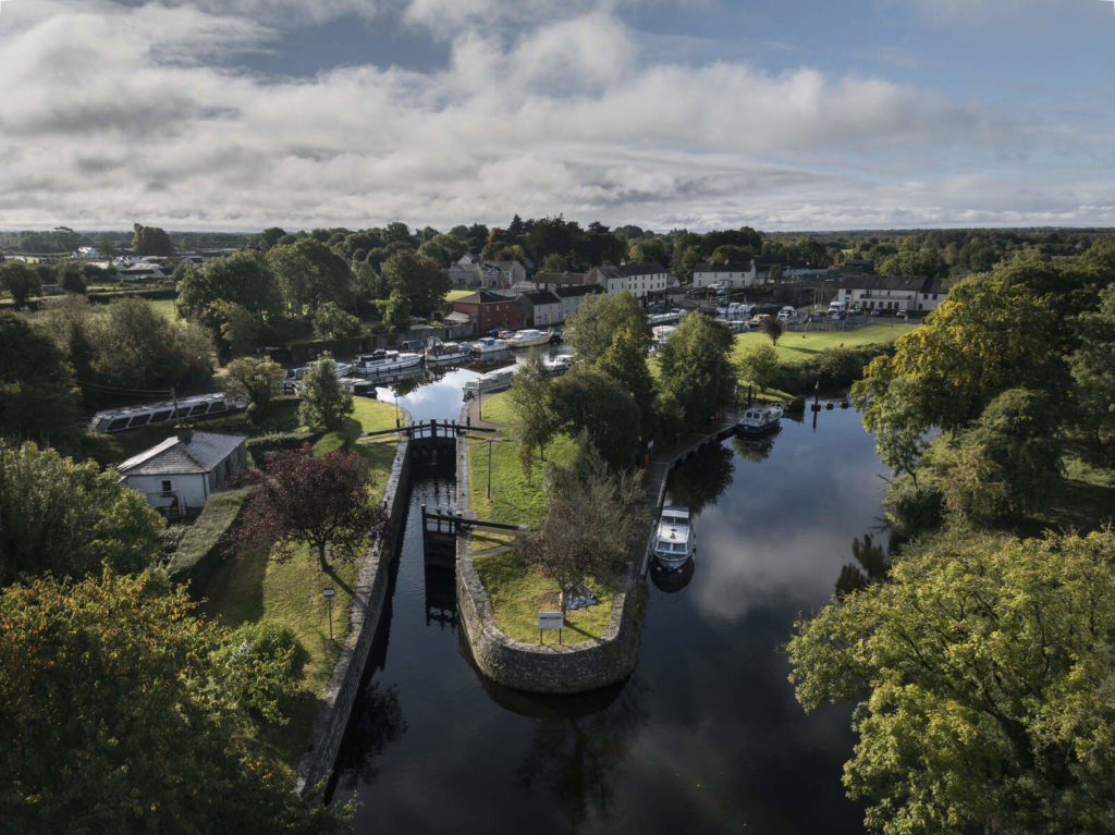 Aerial view of peaceful riverside village with lush greenery, boats, and charming houses under a blue sky.