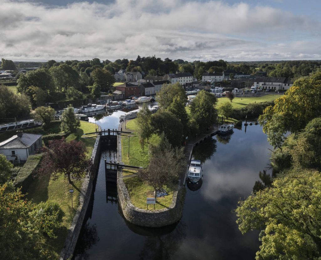 Aerial view of a scenic river lock surrounded by lush greenery and boats in a charming village setting.