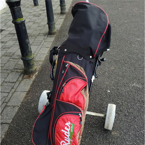 Golf bag with red and black cover on a wheeled cart, parked on a pavement near metal poles.