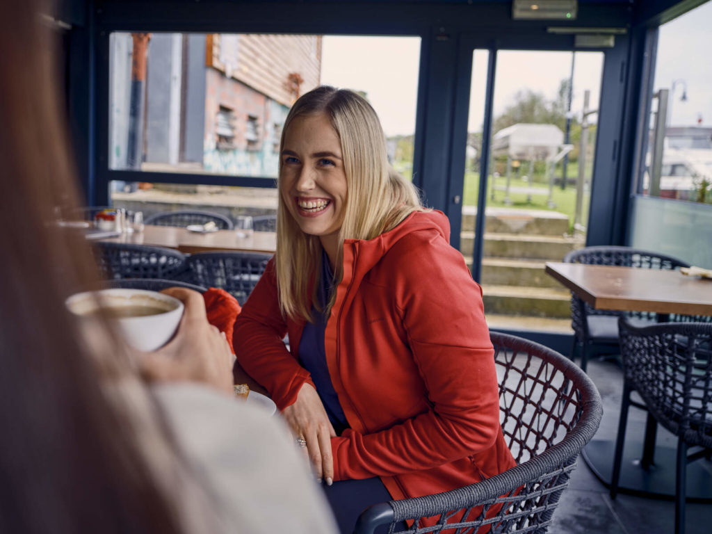 Smiling woman in red jacket enjoying coffee with a friend at a cozy café.