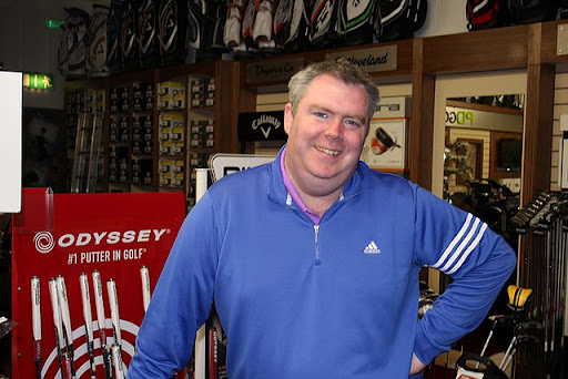 Man in a blue shirt standing in a golf shop with putters and golf equipment in the background.