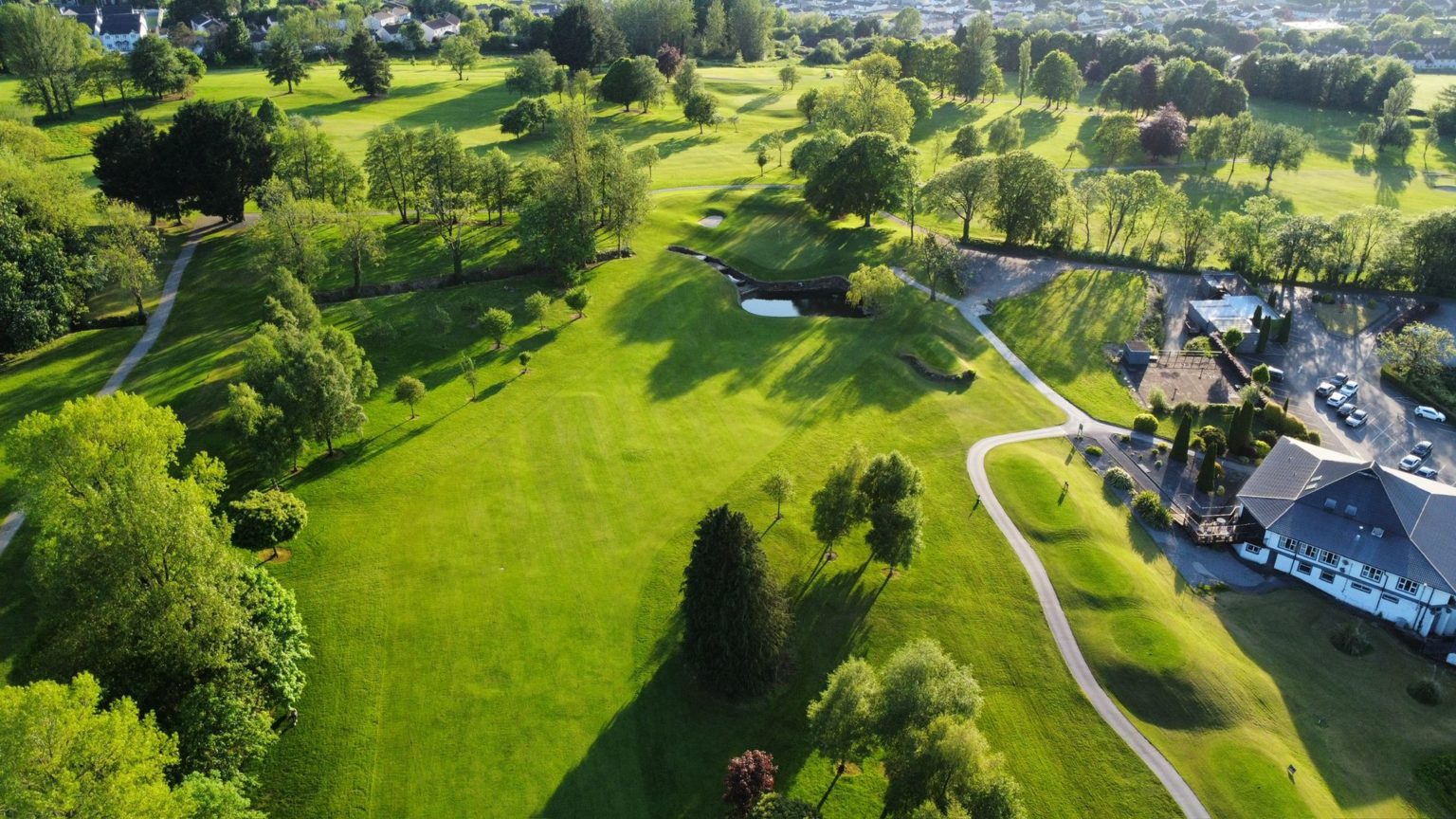 Aerial view of a lush, green golf course with clubhouse and trees under a clear blue sky.