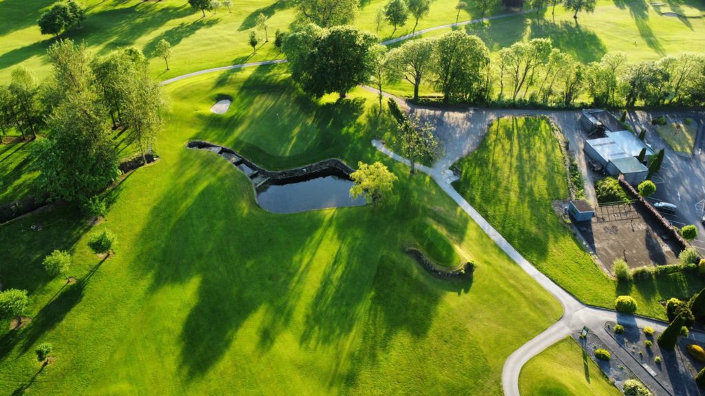 Aerial view of lush golf course landscape with trees, pathways, and a pond on a sunny day.