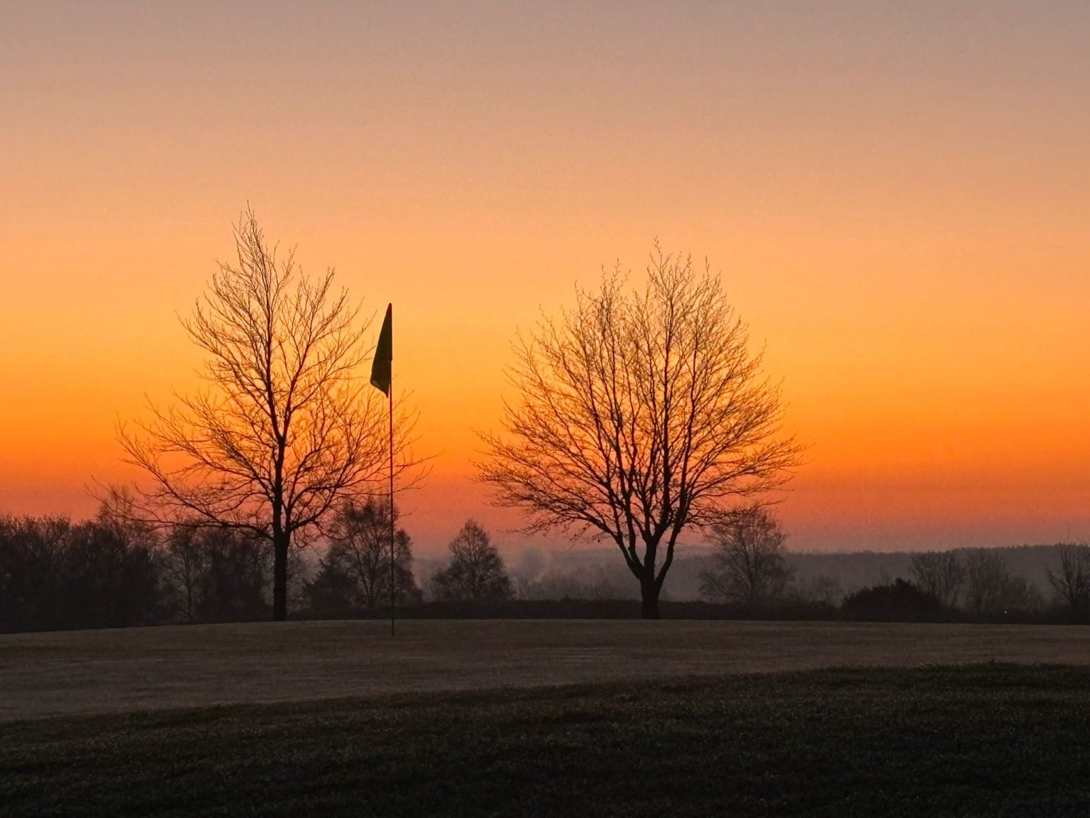Sunrise over a peaceful golf course with silhouette of trees and a flagstick, creating a serene morning scene.