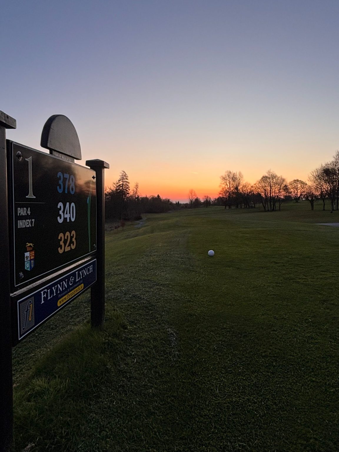 Golf course sunrise, hole 1 sign in foreground, lush green fairway, serene morning sky in the background.