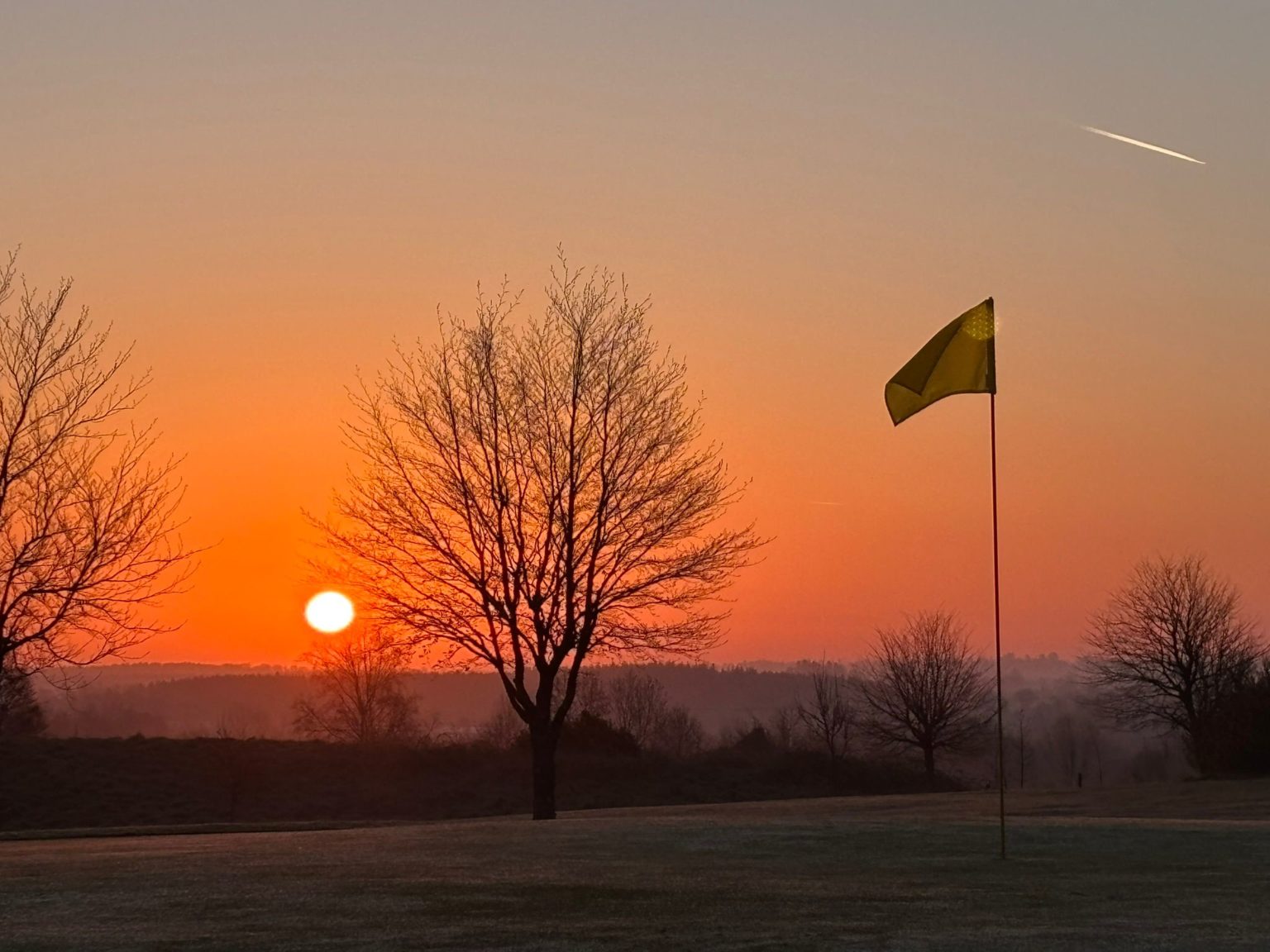 Golf course at sunrise with flag and trees silhouetted against the orange sky.