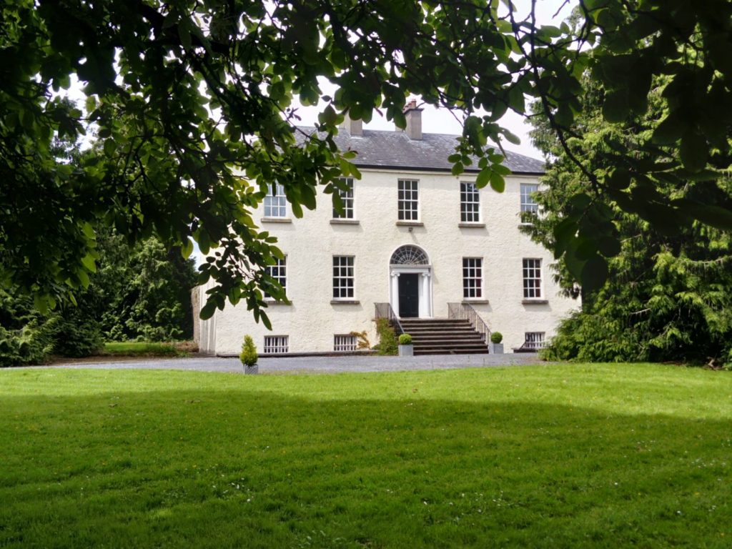 Elegant two-story country house framed by lush green trees and manicured lawn under a partly cloudy sky.