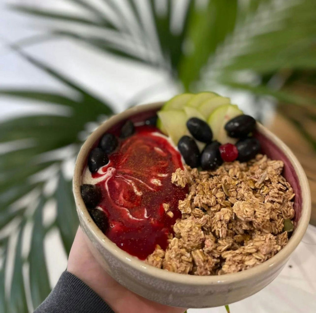 Colorful smoothie bowl with granola, berries, kiwi, and red sauce garnish, held against a leafy background.