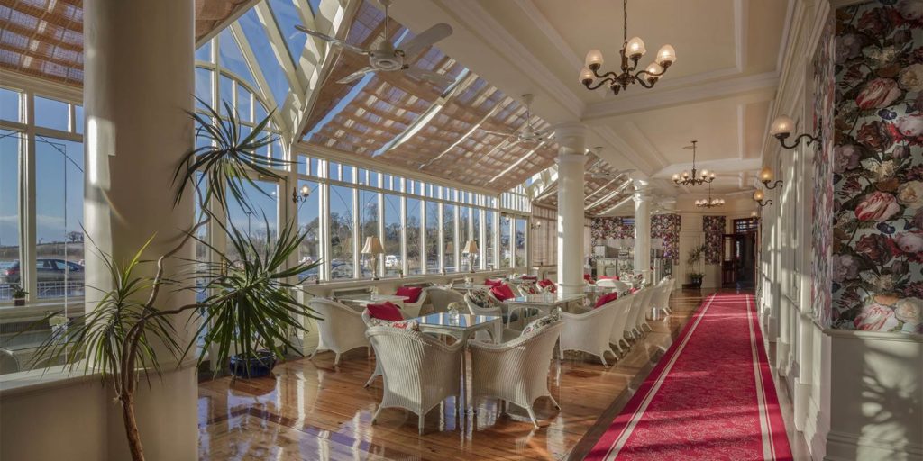 Elegant sunlit restaurant interior with wicker chairs, red accents, and floral wallpaper, featuring large windows and plants.