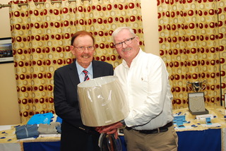 Two men smiling and holding a lamp in a room with a patterned curtain backdrop.