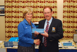 Two people shaking hands during an award ceremony, with a table and patterned curtain in the background.