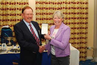 Two people smiling and shaking hands during an award ceremony, with the woman holding a prize against a decorated backdrop.