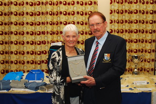 Elderly couple proudly displaying an award in front of a patterned curtain and table with garments and a trophy.