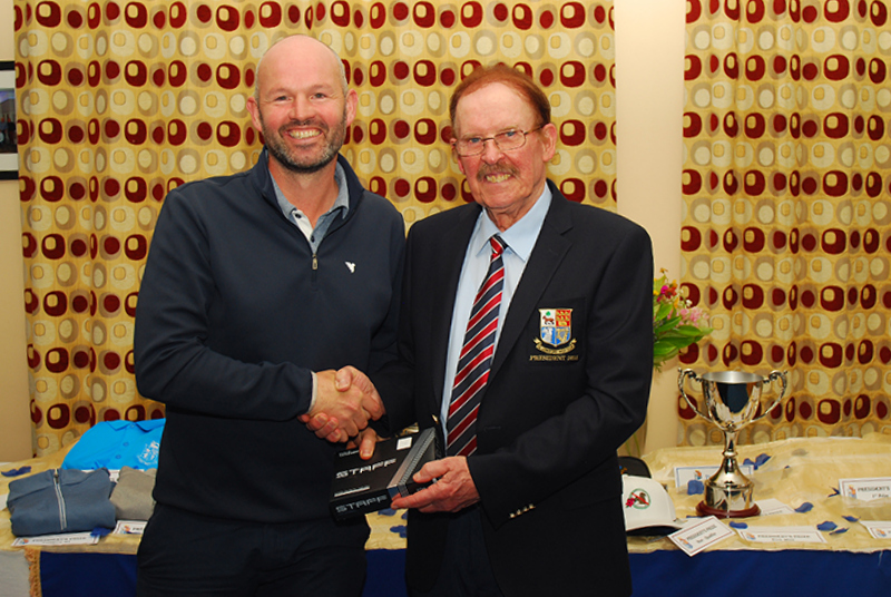 Two men shaking hands at an awards event, with trophies and merchandise in the background.