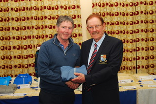 Two men at an event, one presenting a gift, standing in front of a table with fabric items and patterned curtains.