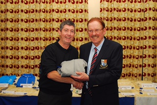 Two men shaking hands and smiling at an award ceremony, with a gift exchange in front of a patterned curtain backdrop.