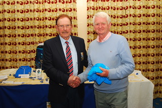 Two men smiling and shaking hands at an indoor event, surrounded by blue and white items on a table.