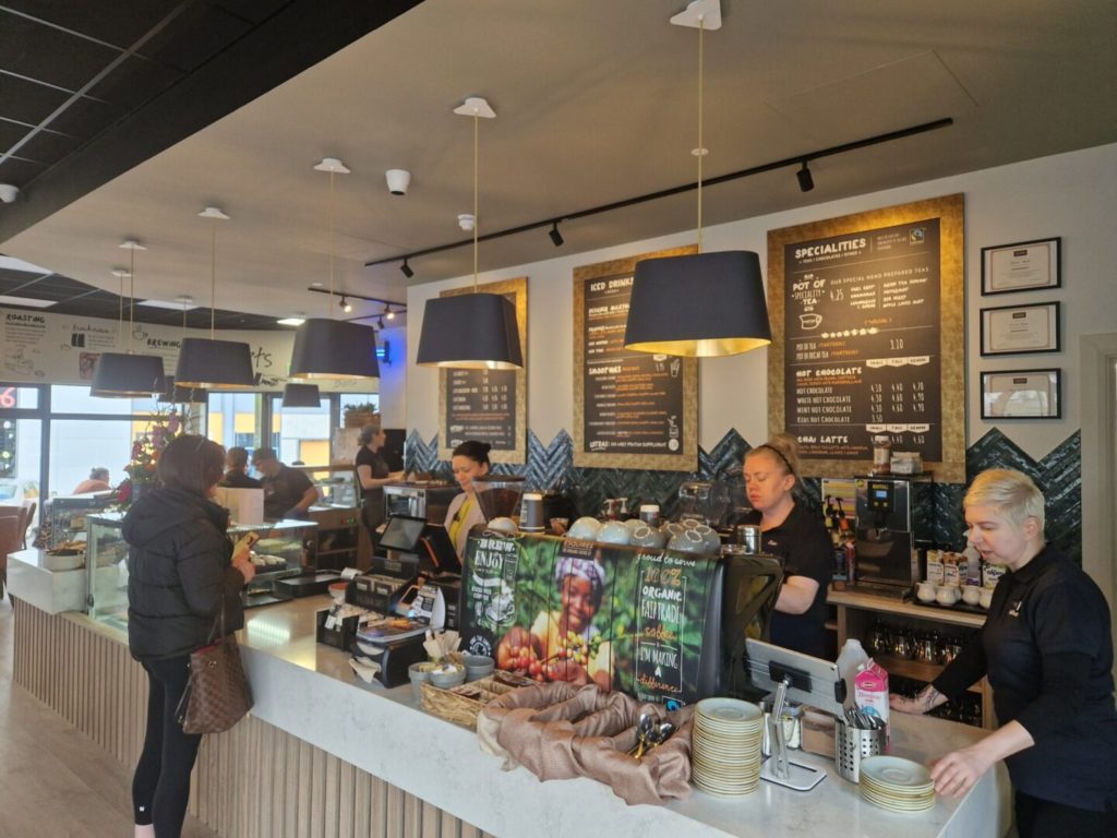 Modern cafe interior with customers and baristas at counter, menus on wall, and stylish lighting fixtures.