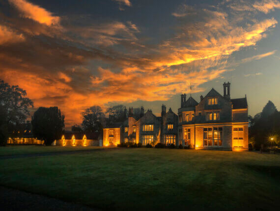 Elegant mansion at sunset with glowing lights, surrounded by lush greenery, beneath a dramatic, colorful sky.