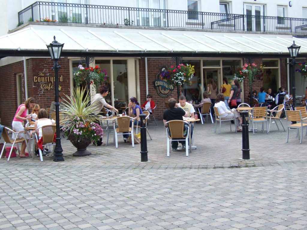 Outdoor seating at a bustling café with people dining and a server attending, surrounded by colorful flowers.