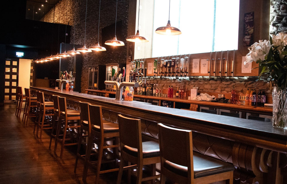 Modern bar interior with long wooden counter, bar stools, hanging lights, and a variety of drinks on display.
