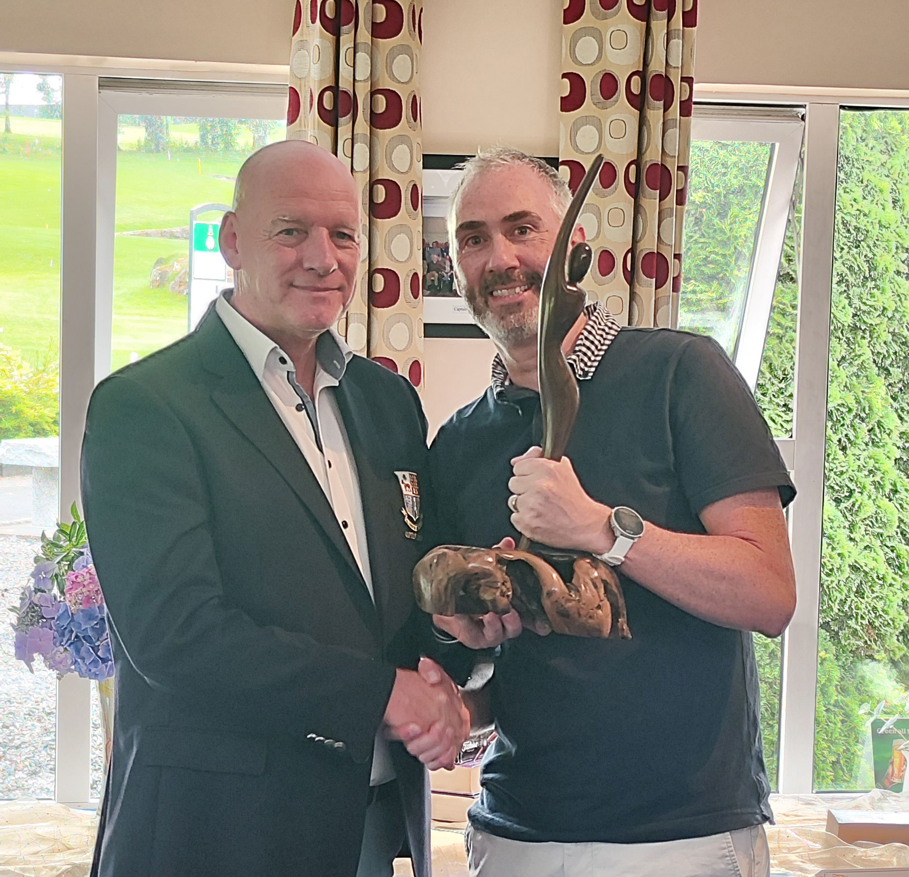 Two men shaking hands; one holds a sculpture, celebrating an achievement indoors.