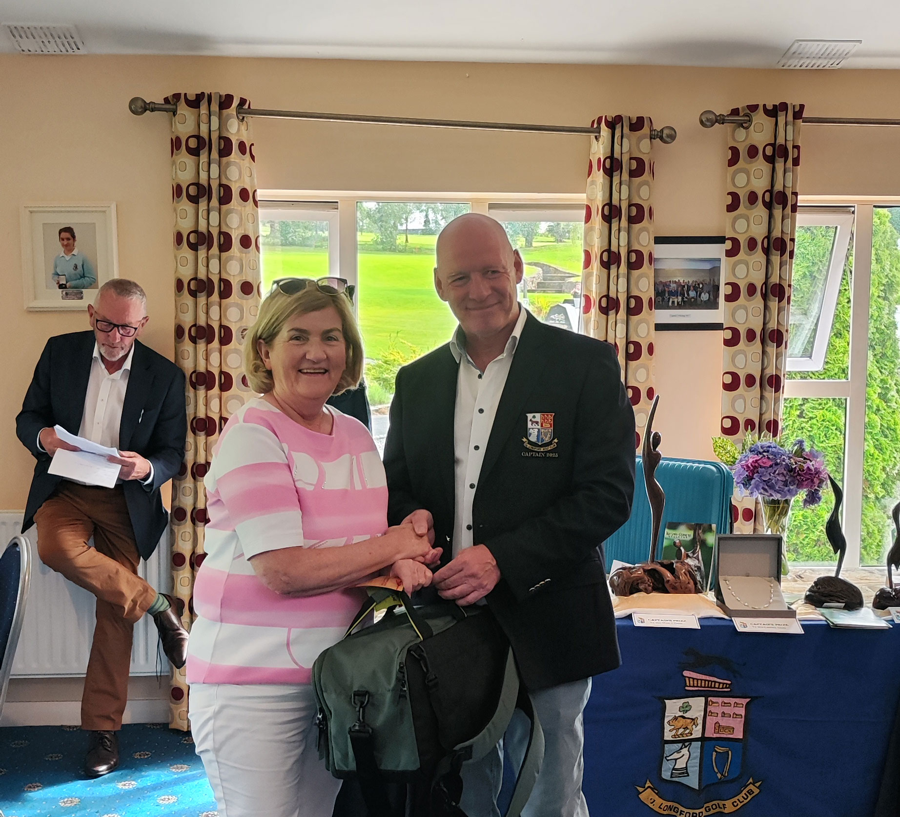 Man and woman shaking hands at an event in a golf club lounge, with awards table in the background.