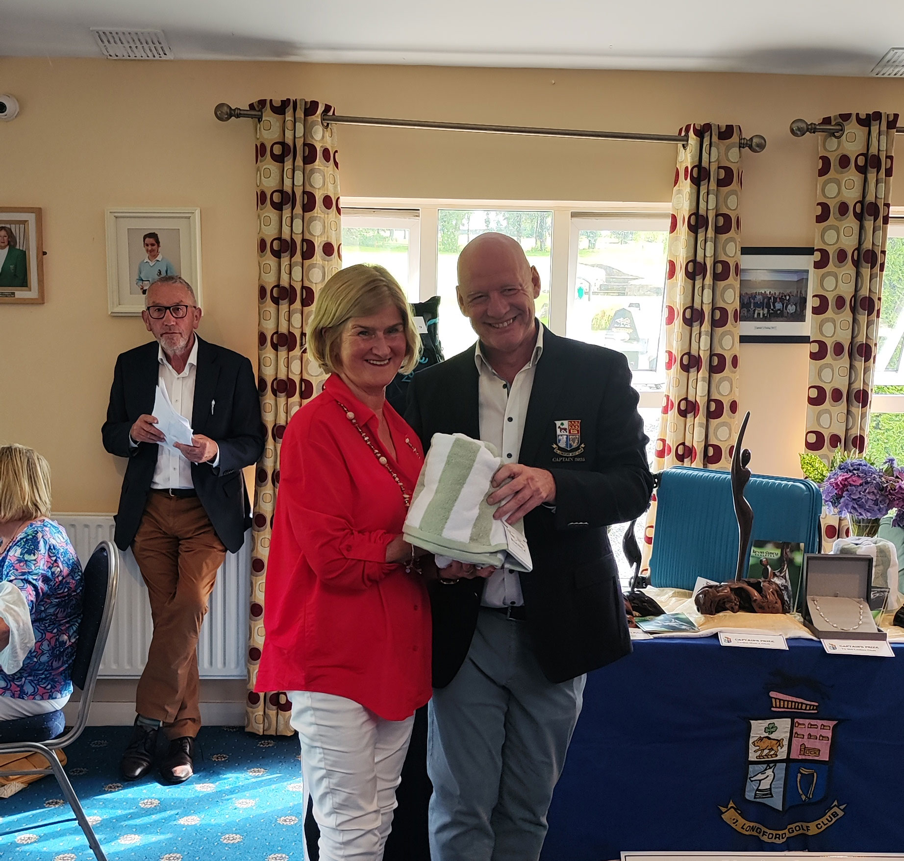 Two people smiling and holding a towel at a social event, with attendees in the background.