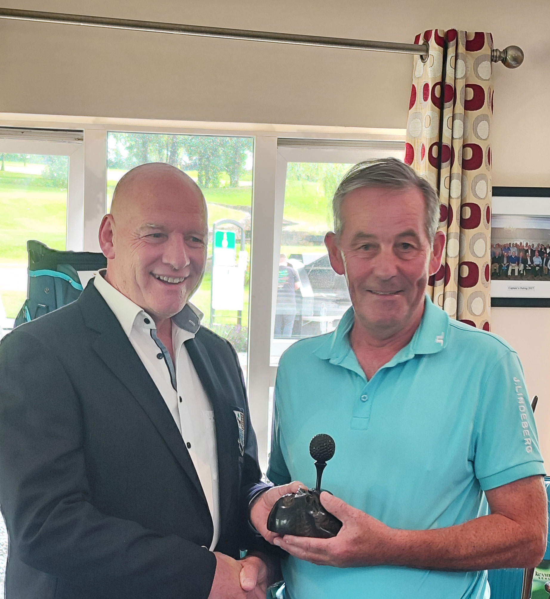 Two men smiling and shaking hands in a room, one holding a trophy or award.