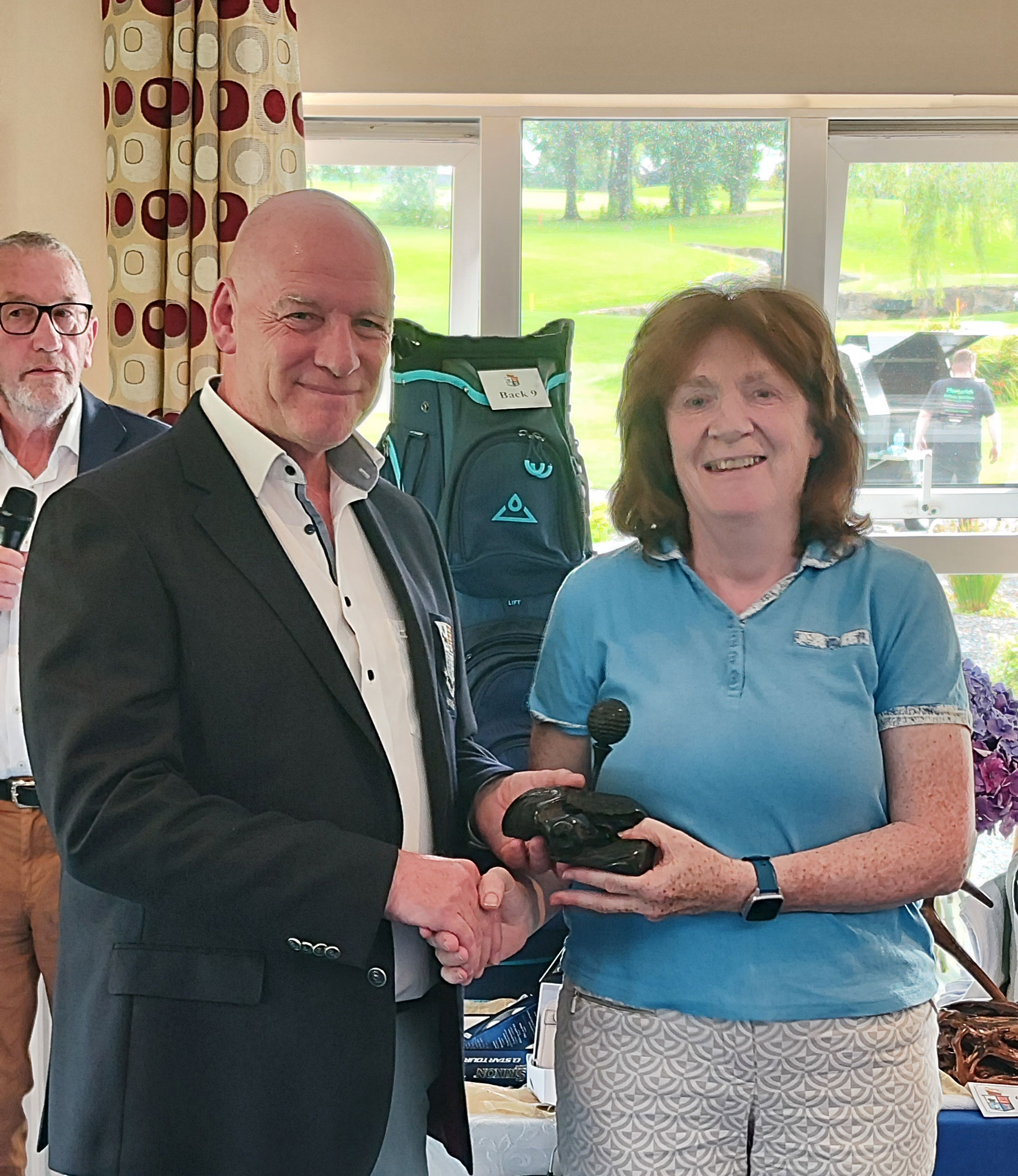 Two people shaking hands at an award ceremony, smiling and holding a trophy indoors.