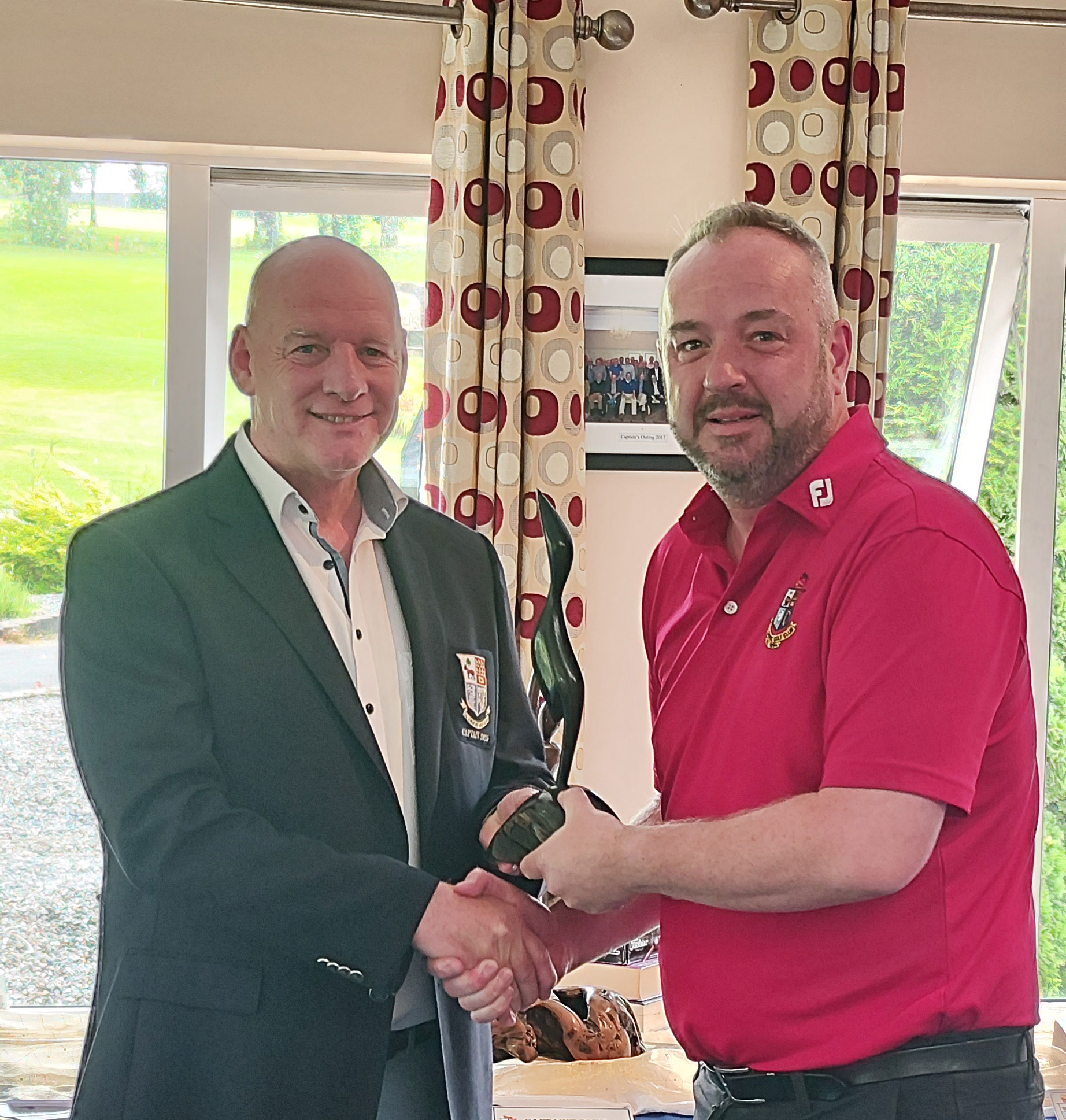 Two men shaking hands, one holding a trophy, standing indoors with a green field visible outside the window.