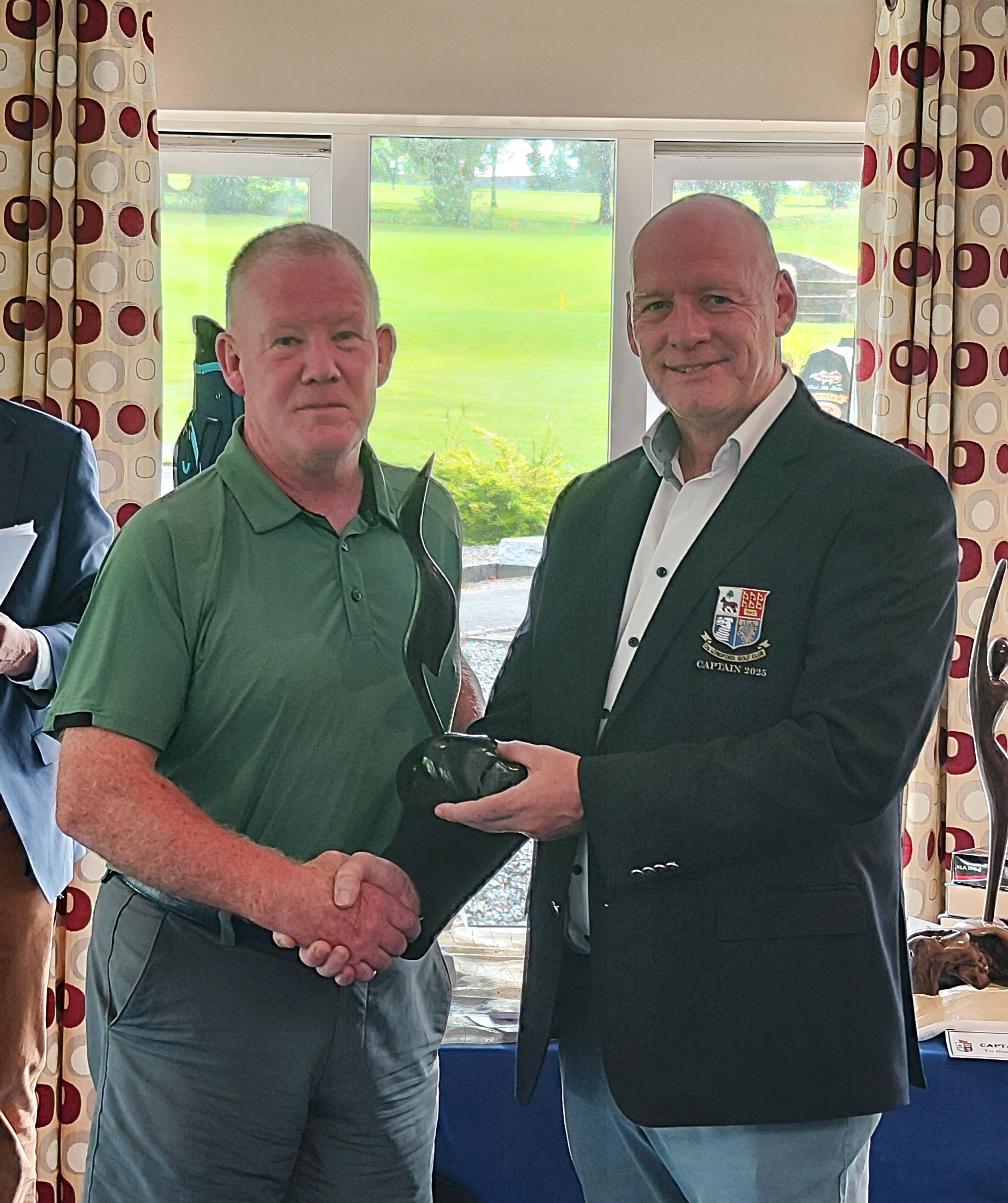 Two men in a formal setting shaking hands while holding a trophy, with a window and curtain backdrop.