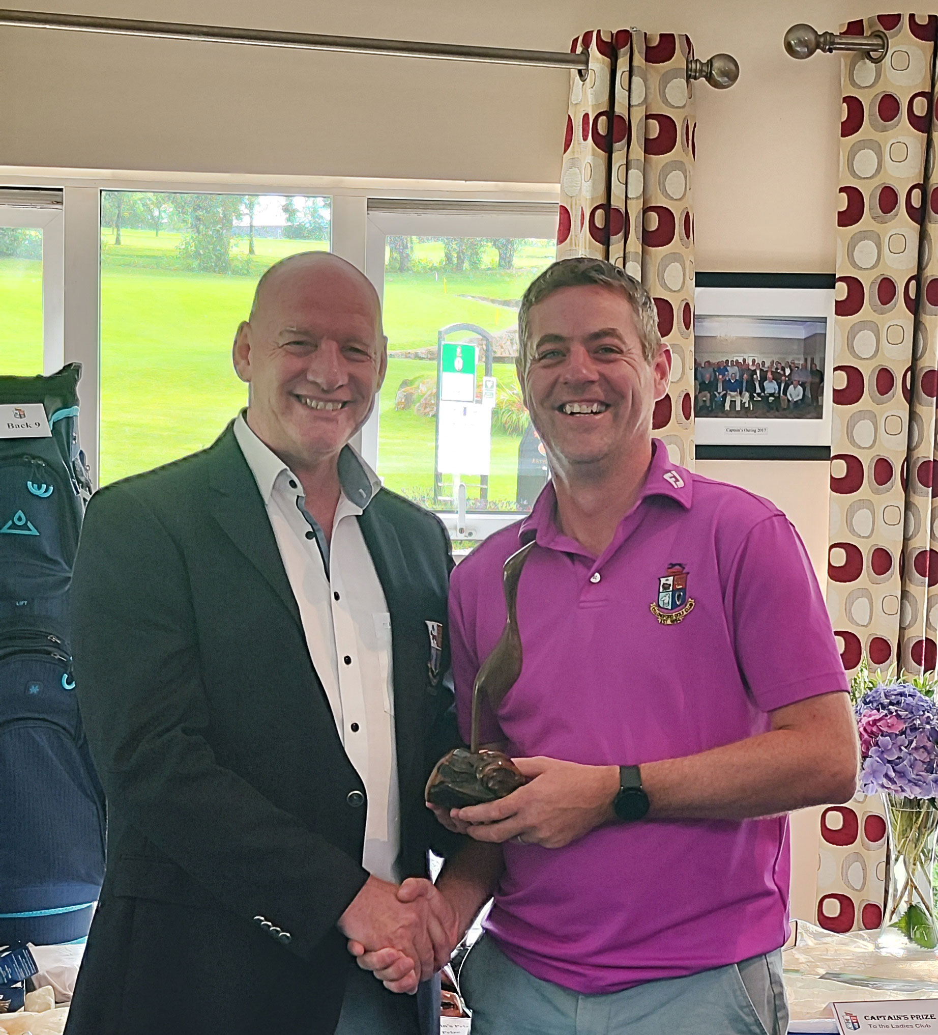 Two men smiling and shaking hands indoors, with one holding a golf trophy. Bright room, golf course view outside.