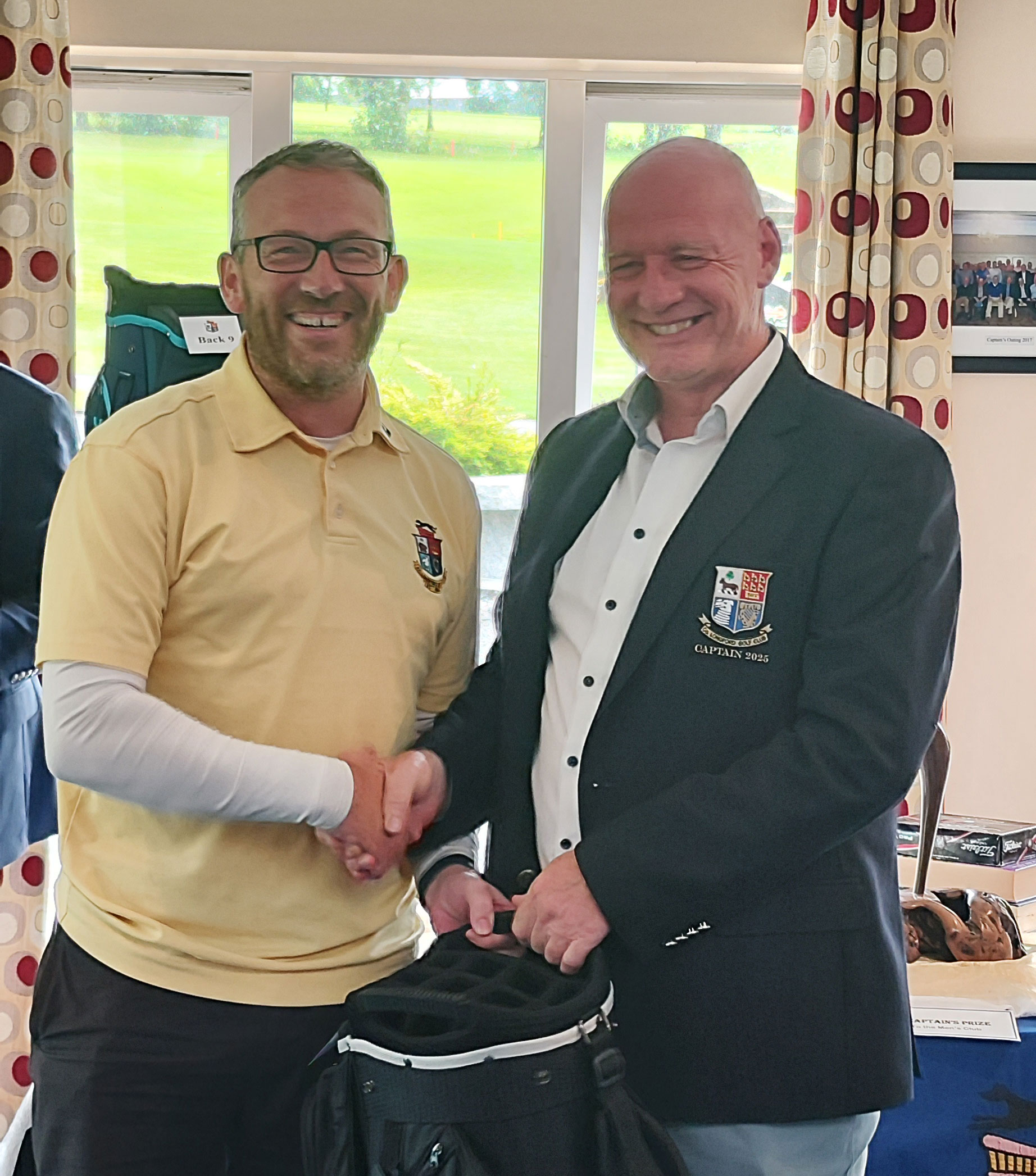Two men shaking hands indoors, one in a yellow shirt, the other in a blazer, standing by a window and golf bag.