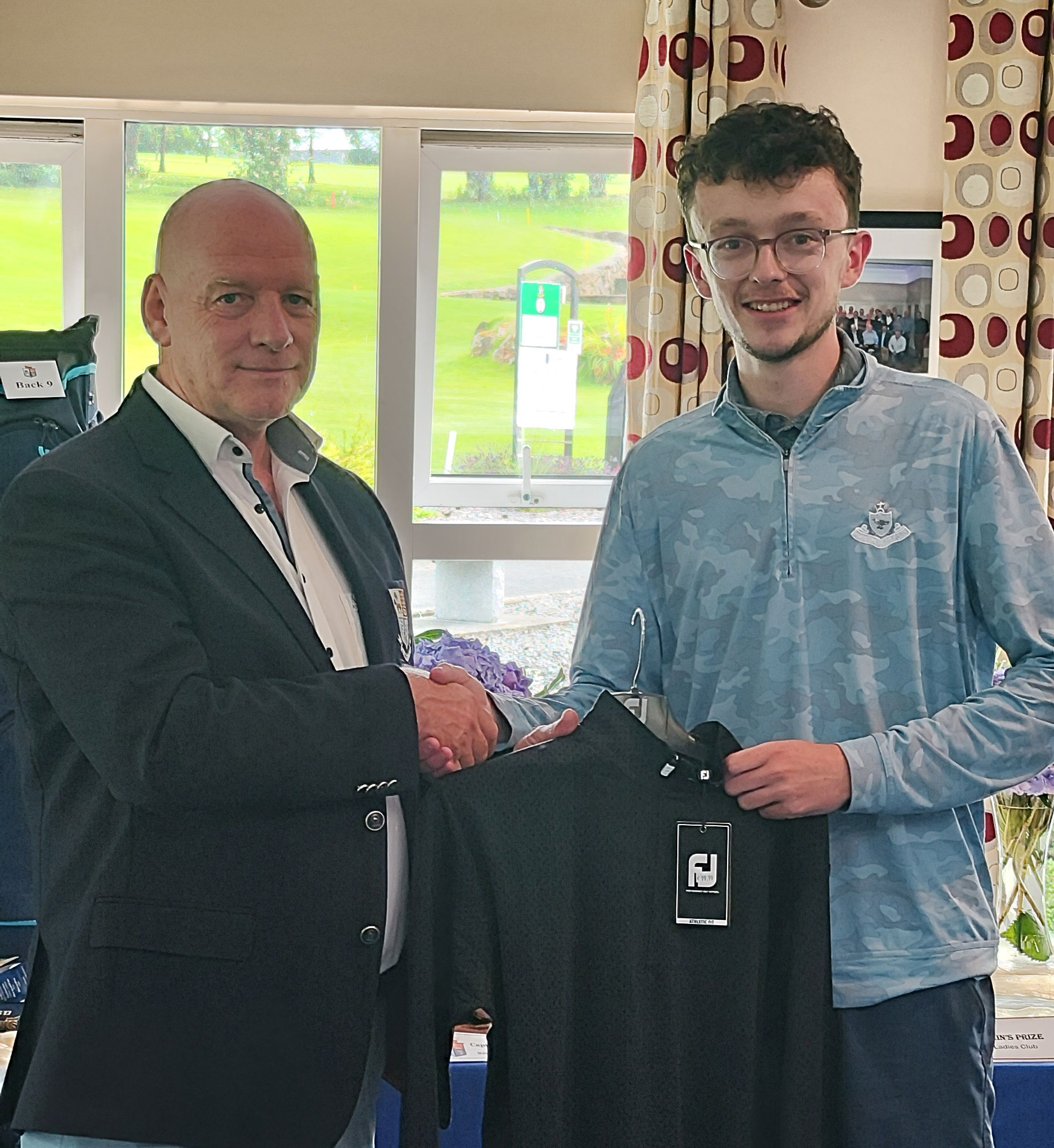 Two men shaking hands indoors, holding a black shirt on a hanger, with golf course visible through windows.