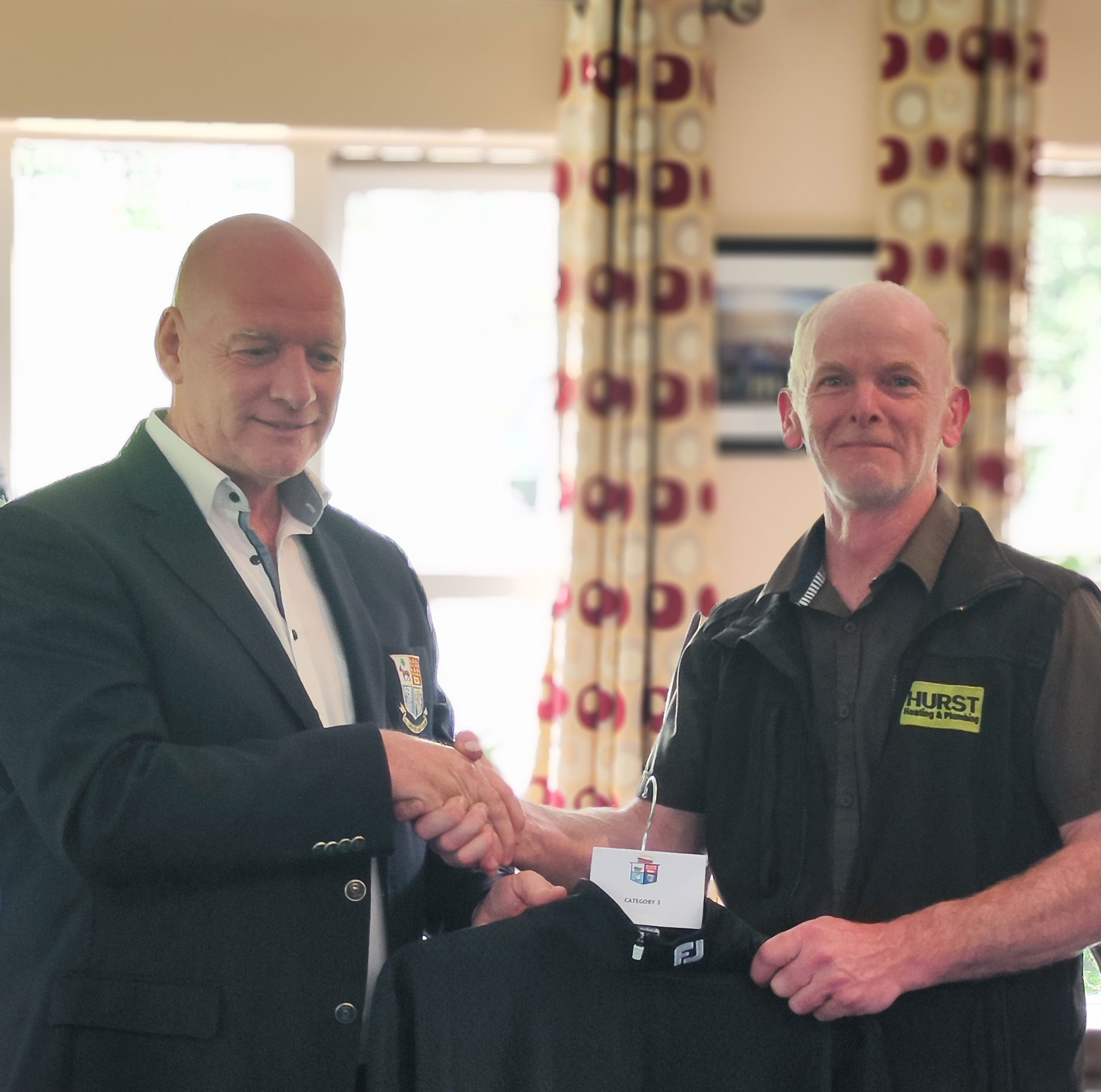 Two men shaking hands indoors, one holding a jacket with a name tag labeled Category 1.