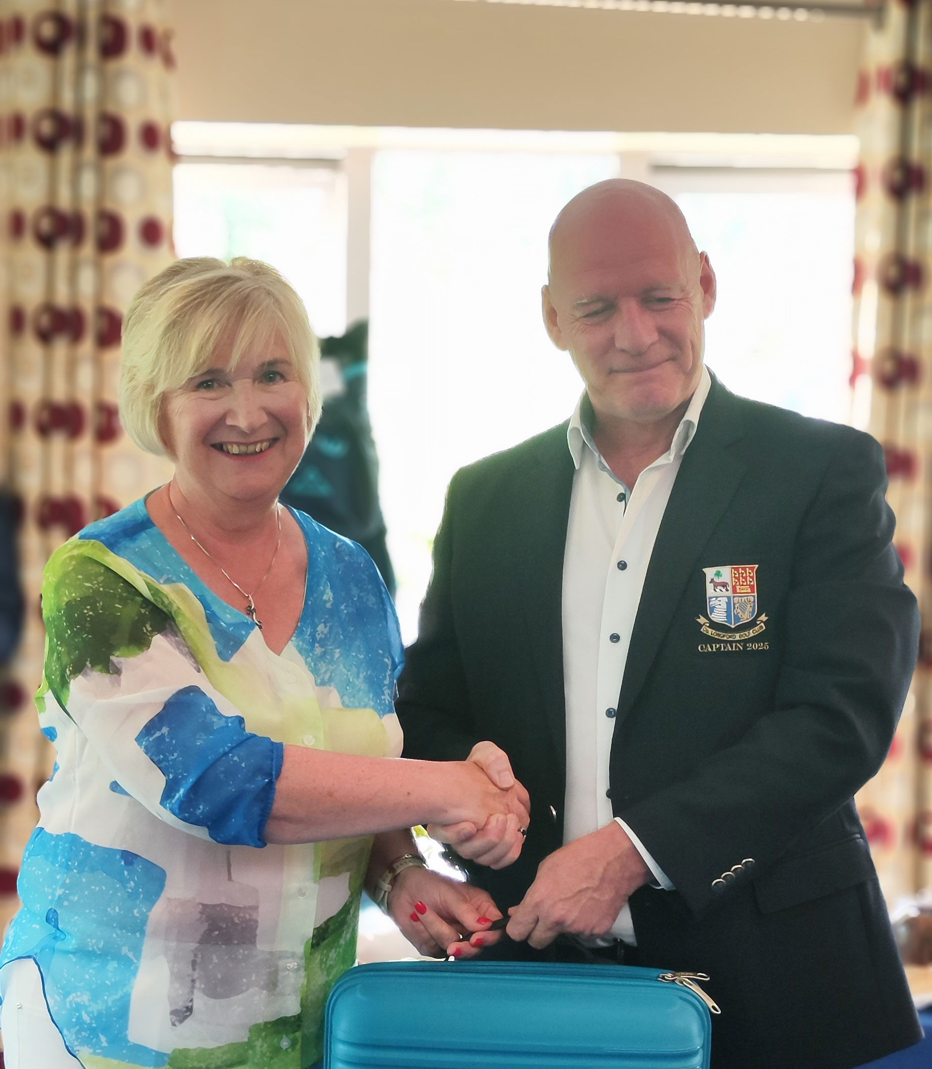 Man and woman shaking hands in a room, smiling, with a suitcase in front. He's wearing a blazer with a Captain 2023 crest.