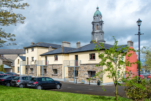 Historic stone building with a tall clock tower under a cloudy sky, cars parked in front, and trees nearby.