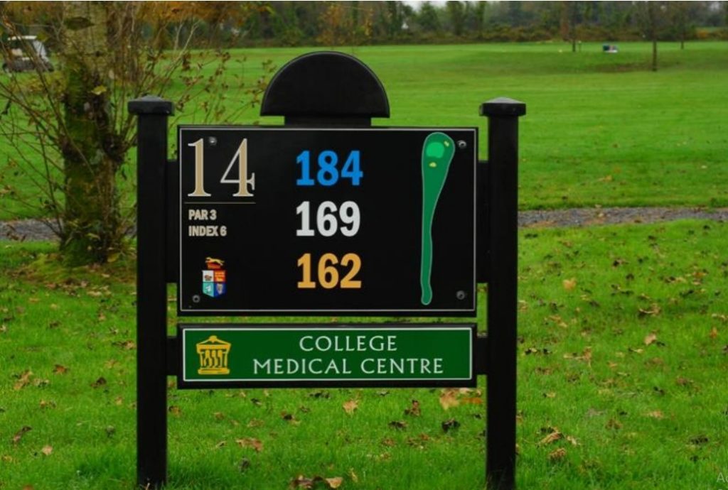 Golf course sign for hole 14, showing distances for par 3 and College Medical Centre logo in a grassy field.