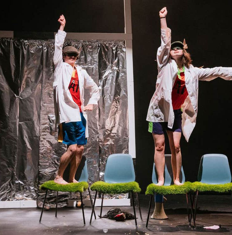 Two performers in lab coats dance energetically on chairs in a theatrical play.