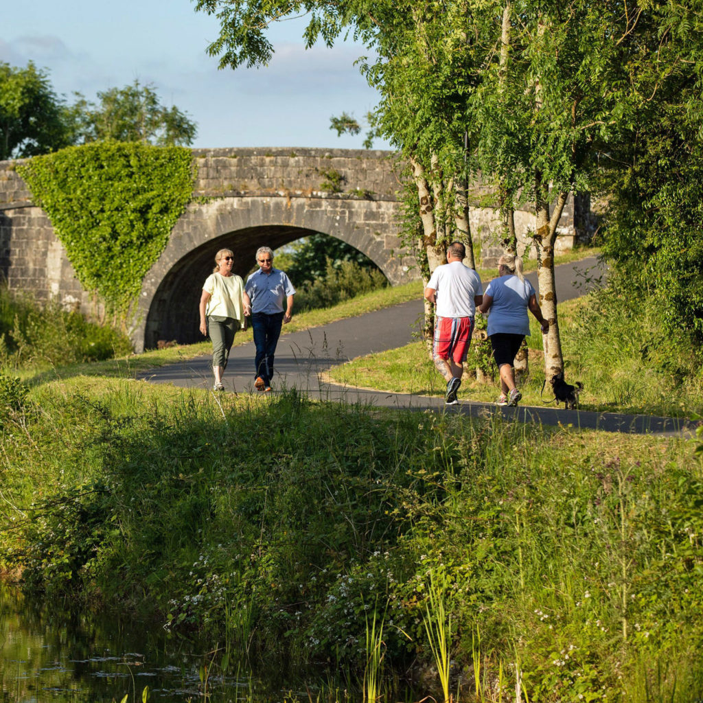 People walking on scenic path near an ivy-covered stone bridge, surrounded by lush greenery and bright summer light.