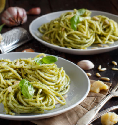 Two plates of spaghetti with green pesto sauce, garnished with basil leaves and parmesan, on a dark wooden table.
